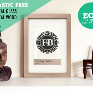 May include: A light wood picture frame with a white mount and a black and white logo that says "Farrow & Ball Paint & Paper" and "Setting Plaster No. 211". The frame is on a white shelf with a wooden statue and a small wooden chair.