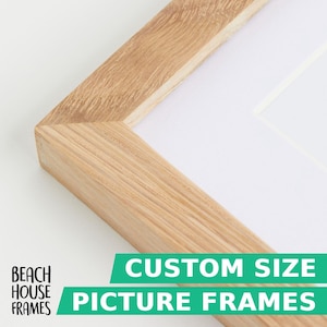 May include: A light brown wooden picture frame with a visible natural grain pattern and a white mat board border. The frame features mitered corners and a clean, simple construction. Text on the image reads: BEACH HOUSE FRAMES CUSTOM SIZE PICTURE FRAMES.