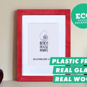 Könnte beinhalten: Ein rechteckiger roter Holzbilderrahmen mit weißem Passepartout. In der Mitte befindet sich ein Druck mit einem Nautilus-Muschel-Symbol und dem Text "BEACH HOUSE FRAMES" sowie "HANDMADE IN". Grüne Etiketten besagen "ECO FRIENDLY PACKAGING", "PLASTIC FREE", "REAL GLASS" und "REAL WOOD".