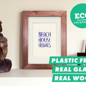 Könnte beinhalten: Ein Holzbildrahmen mit einem weißen Passepartout und dem Text "Beach House Frames" in Lila. Der Rahmen steht auf einem weißen Regal mit einer Holzstatue und einem Holzstuhl. Der Text "Plastic Free, Real Glass, Real Wood" ist in Weiß auf grünem Hintergrund geschrieben.