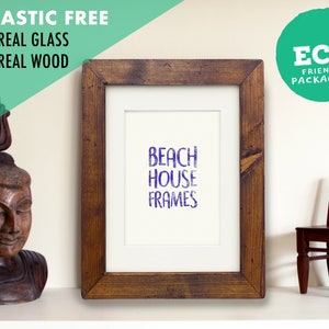 May include: A rustic wooden picture frame with a white mat and a purple inscription that reads "Beach House Frames". The frame is sitting on a white surface with a wooden statue and a small wooden chair.
