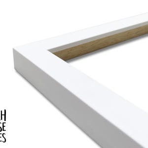 May include: A corner section of a rectangular picture frame with a smooth matte white finish on the exterior and a natural light brown wood grain visible on the inner edge. The text BEACH HOUSE FRAMES is printed in black in the bottom left corner.