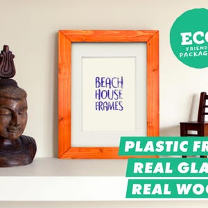 Peut inclure: Un cadre photo rectangulaire en bois avec une finition orange-brun naturel et un passe-partout blanc. Le cadre contient un insert blanc avec le texte bleu BEACH HOUSE FRAMES. Un badge circulaire vert indique ECO FRIENDLY PACKAGING.