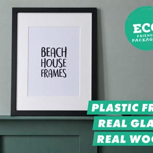 Op de afbeelding: Een zwarte houten fotolijst met een witte passe-partout en een witte print met de zwarte tekst BEACH HOUSE FRAMES. De lijst is voorzien van echt glas en is verpakt in milieuvriendelijke, plasticvrije materialen, zoals aangegeven op het label.