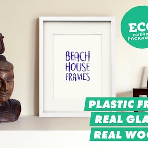 Könnte beinhalten: Weißer Bilderrahmen mit einem weißen Passepartout und einem violetten Druck, auf dem "Beach House Frames" steht. Der Rahmen steht auf einem weißen Regal mit einer Holzstatue links und einem Holzstuhl rechts. Der Text "Plastic Free, Real Glass, Real Wood" befindet sich unter dem Rahmen.
