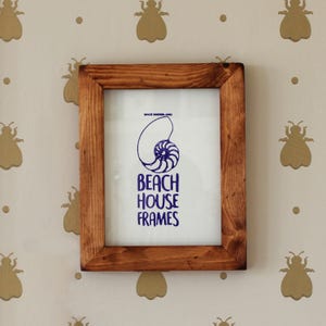 Puede incluir: Un marco de fotos de madera con un acabado marrón claro. El marco está colgado en una pared con un fondo blanco y un patrón de abejas doradas. El marco contiene una impresión con una concha de nautilus y el texto "Beach House Frames".