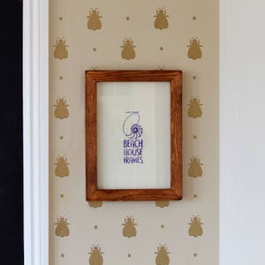May include: A brown wooden picture frame with a white mount and a blue and white print inside. The print features a seashell and the text "Beach House Frames". The frame is hanging on a wall with a beige background and a pattern of gold bee silhouettes.