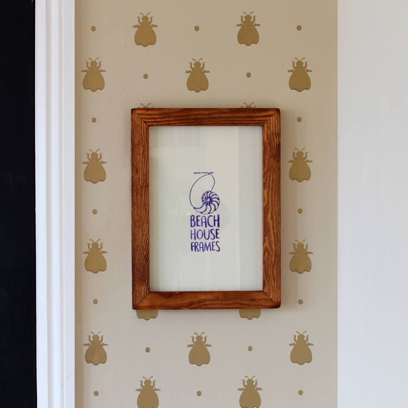 May include: A brown wooden picture frame with a white mount and a blue and white print inside. The print features a seashell and the text "Beach House Frames". The frame is hanging on a wall with a beige background and a pattern of gold bee silhouettes.