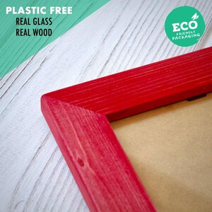 Peut inclure: Un coin de cadre photo en bois rouge vif avec une texture de grain de bois visible. Le cadre présente un panneau arrière beige. Le texte sur l'image indique : PLASTIC FREE, REAL GLASS, REAL WOOD, et un badge circulaire vert ECO FRIENDLY PACKAGING.