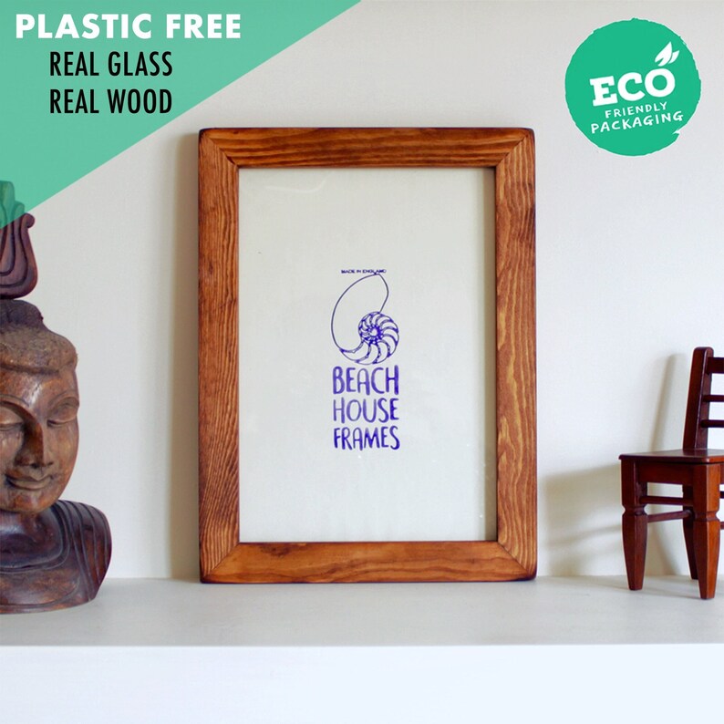 May include: A wooden picture frame with a light brown stain. The frame is empty and has a white mount. The frame is sitting on a white shelf. The text "Beach House Frames" is printed in purple on the mount. A small wooden chair is in the background.