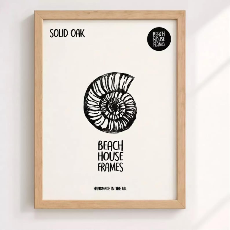 The image is a framed artwork featuring a black and white illustration of a spiral shell or nautilus shell. The text on the frame reads "SOLID OAK" and "BEACH HOUSE FRAMES" along with "HANDMADE IN THE UK".