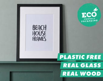 Könnte beinhalten: Ein schwarzer Holzbilderrahmen mit weißem Passepartout und dem Text BEACH HOUSE FRAMES in schwarzer Schrift. Ein grünes kreisförmiges Abzeichen in der Ecke zeigt den Text ECO FRIENDLY PACKAGING, PLASTIC FREE, REAL GLASS, REAL WOOD.