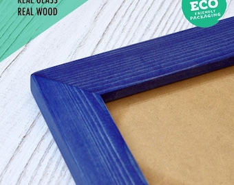 Può includere: Una cornice portafoto in legno blu con una texture venata del legno visibile. La cornice presenta un pannello posteriore marrone. Il testo sull'immagine recita: PLASTIC FREE, REAL GLASS, REAL WOOD, e un badge circolare verde ECO FRIENDLY PACKAGING.