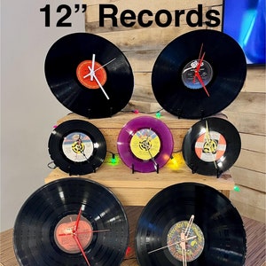 Puede incluir: Siete discos de vinilo de 30 cm convertidos en relojes. Los discos son negros con varias portadas de álbum visibles. Los relojes tienen manecillas blancas y rojas. Los discos se muestran en una superficie de madera con luces navideñas de colores.