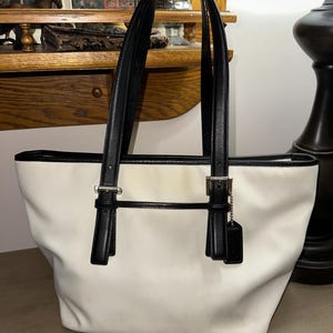 May include: Cream-colored tote bag with black leather handles and trim. The bag features silver-tone hardware and a small black tag. The bag is made of a canvas-like material and has a rectangular shape.