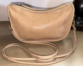 May include: A light brown leather shoulder bag with a curved shape and a long strap. The bag has a simple design and a worn look.