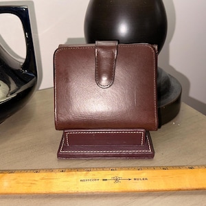 Op de afbeelding: Een bruine leren portemonnee met een klep sluiting en een bordeauxrood interieur. De portemonnee staat rechtop op een tafel.
