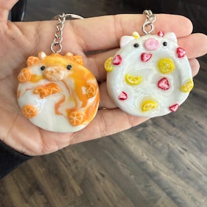 May include: Two handmade keychains. One is orange and white, resembling a citrus fruit. The other is white with red and yellow fruit slices. Both have silver key rings.