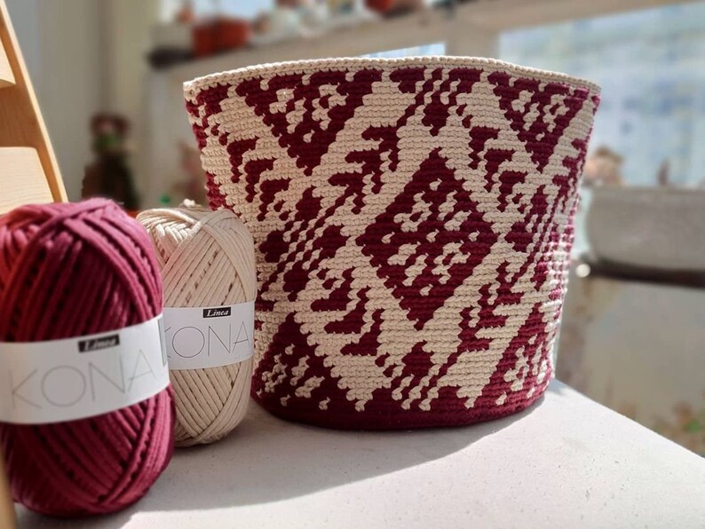 May include: Two balls of yarn, one burgundy and one beige, with the label "Lina Kona" on each. A crocheted basket with a geometric pattern in burgundy and beige.