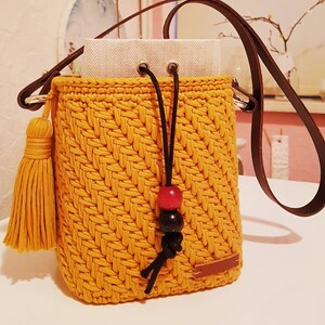 May include: A mustard yellow crocheted bucket bag with a brown leather strap, black drawstring, and a yellow tassel. The bag has a red and black bead accent.