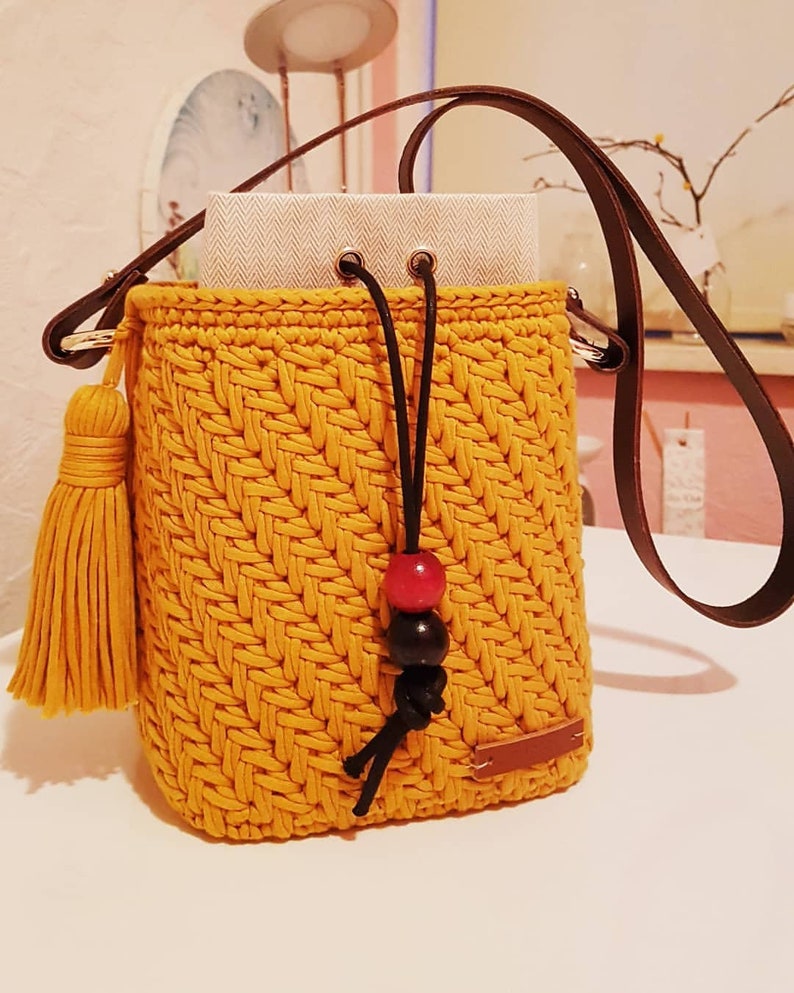 May include: A mustard yellow crocheted bucket bag with a brown leather strap, black drawstring, and a yellow tassel. The bag has a red and black bead accent.