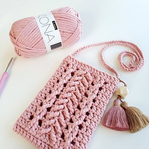 May include: A pink crocheted bag with a long strap and two tassels. The bag is made with thick yarn and has a woven pattern. The yarn is in a ball next to the bag.