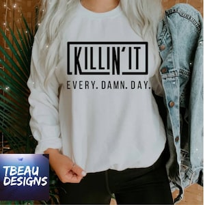 May include: White sweatshirt with the text "KILLIN' IT EVERY. DAMN. DAY." in a black rectangular box. The sweatshirt is long-sleeved and has a crew neck. A denim jacket is draped over the shoulder. The TBEAU DESIGNS logo is in the bottom left corner.