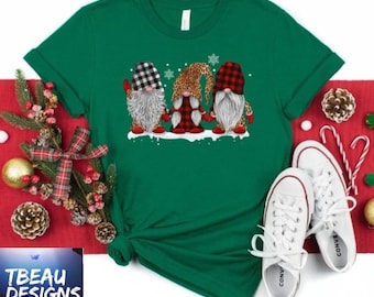 May include: A green t-shirt featuring three gnome characters with plaid, leopard, and white beard designs. The gnomes are standing on a snowy base. The shirt is paired with white sneakers and holiday decorations. The text "TBEAU DESIGNS" is in the lower left corner.