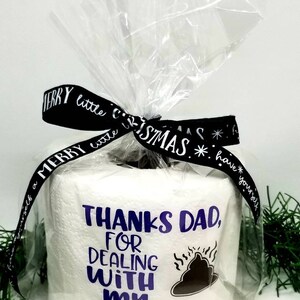May include: A roll of white toilet paper wrapped in clear cellophane, tied with a black ribbon that says "Merry Little Christmas." The toilet paper has the text "Thanks Dad, for dealing with my" and a graphic.