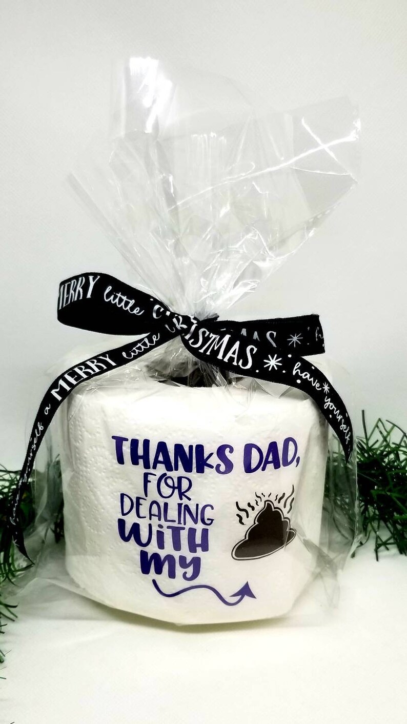 May include: A roll of white toilet paper wrapped in clear cellophane, tied with a black ribbon that says "Merry Little Christmas." The toilet paper has the text "Thanks Dad, for dealing with my" and a graphic.