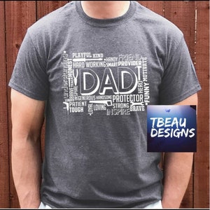 May include: Dark gray t-shirt with the word "DAD" in large white letters. Surrounding the word are smaller words like "Protector," "Loving," and "Brave." The shirt also features the text "TBEAU DESIGNS" in a small box.