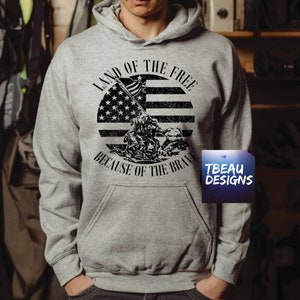 May include: A gray hooded sweatshirt with a front graphic featuring an American flag design and the text "LAND OF THE FREE BECAUSE OF THE BRAVE." The design is in black and white, with a distressed look. The sweatshirt has a front pocket.