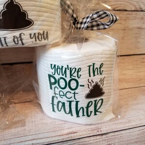 May include: White toilet paper rolls with humorous text and a poop graphic. One roll reads "You're the POO-fect father" in green. Another roll has a similar graphic and text. The rolls are wrapped in clear cellophane and tied with a black and white checkered ribbon.