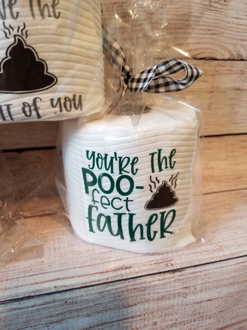 May include: White toilet paper rolls with humorous text and a poop graphic. One roll reads "You're the POO-fect father" in green. Another roll has a similar graphic and text. The rolls are wrapped in clear cellophane and tied with a black and white checkered ribbon.