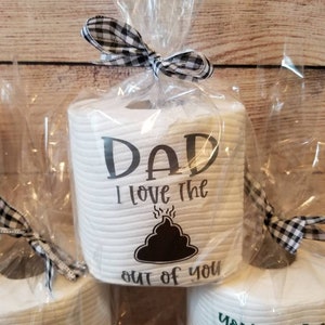 May include: White toilet paper rolls with humorous text, wrapped in clear cellophane and tied with a black and white checkered bow. The text on the front of the roll says "DAD I love the out of you" with a poop emoji.