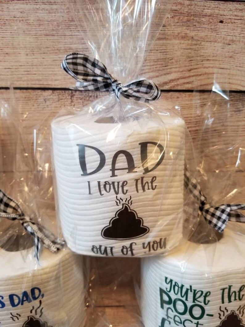 May include: White toilet paper rolls with humorous text, wrapped in clear cellophane and tied with a black and white checkered bow. The text on the front of the roll says "DAD I love the out of you" with a poop emoji.