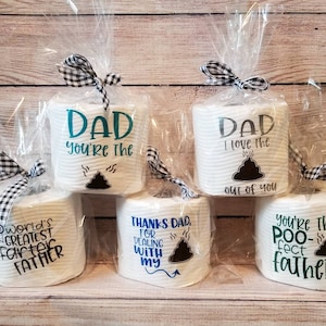 May include: Several rolls of white toilet paper are wrapped in clear cellophane and tied with black and white checkered bows. Each roll has a different message printed on it, including "Dad, you're the..." and "World's Greatest Father."