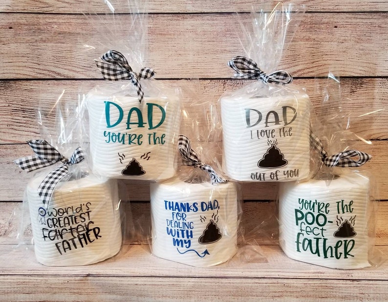 May include: Several rolls of white toilet paper are wrapped in clear cellophane and tied with black and white checkered bows. Each roll has a different message printed on it, including "Dad, you're the..." and "World's Greatest Father."