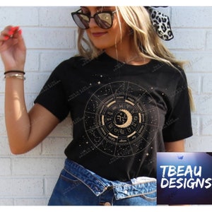 K&ouml;nnte beinhalten: Schwarzes T-Shirt mit einem goldenen Tierkreisrad-Design, einschlie&szlig;lich Sternbildern und astrologischen Symbolen. Das T-Shirt hat einen Rundhalsausschnitt und kurze &Auml;rmel. Der Text "THIS IS A LDC ORIGINAL DESIGN" ist auf dem T-Shirt aufgedruckt. Das TBEAU DESIGNS-Logo befindet sich unten rechts.