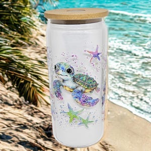 May include: A clear glass tumbler with a bamboo lid, featuring a colorful illustration of a sea turtle and starfish. The design includes shades of blue, green, and purple, with glitter accents. The tumbler is cylindrical and appears to be approximately 6 inches tall.