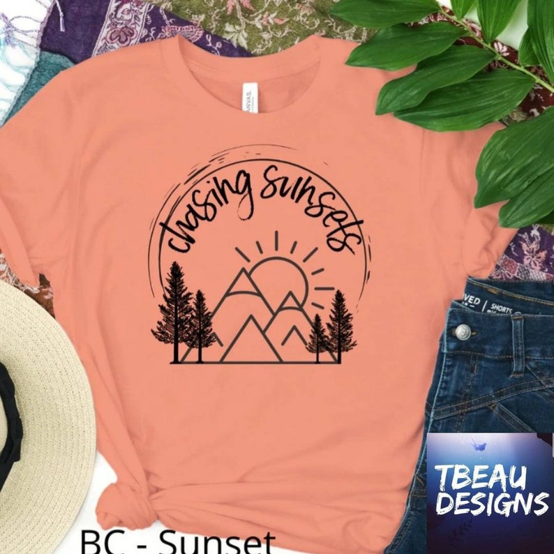 May include: Peach-colored t-shirt with a black graphic design featuring the text "Chasing Sunsets" above a mountain and sun illustration. The shirt has short sleeves and a crew neck. The brand name "TBEAU DESIGNS" is visible on the bottom right.
