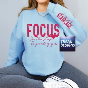 May include: Light blue sweatshirt with the word "FOCUS" in distressed pink lettering. Below the word "FOCUS" is the phrase "On the step In front of you" and a pink heart. The sleeve reads "Not the whole STAIRCASE" in pink. The brand name "TBEAU DESIGNS" is in the lower right corner.