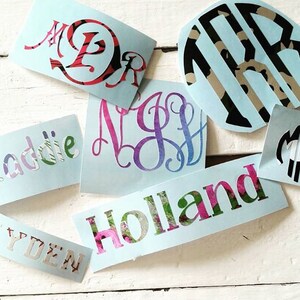 Op de afbeelding: Een verzameling lichtblauwe monogram- en naamstickers. De stickers hebben verschillende lettertypen en ontwerpen, waaronder bloemen- en patroonvullingen. De namen "Holland" en "Addie" zijn zichtbaar, samen met monograminitialen.