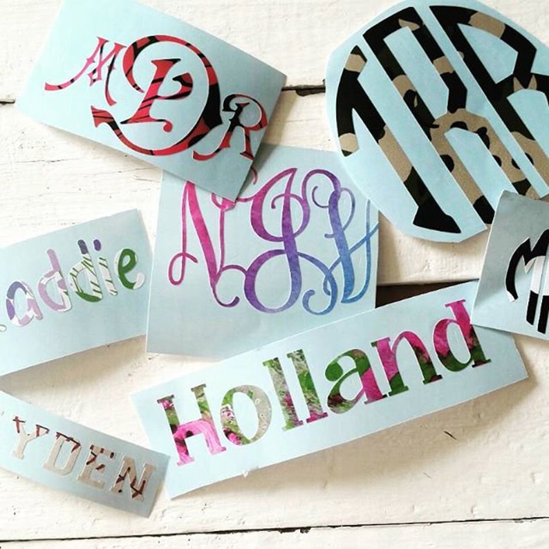 Op de afbeelding: Een verzameling lichtblauwe monogram- en naamstickers. De stickers hebben verschillende lettertypen en ontwerpen, waaronder bloemen- en patroonvullingen. De namen "Holland" en "Addie" zijn zichtbaar, samen met monograminitialen.