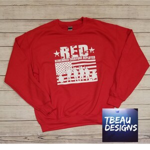 May include: A red crew neck sweatshirt featuring the word "RED" in white, with the text "REMEMBER EVERYONE DEPLOYED." A white graphic of the American flag and soldier silhouettes is below the text. The sweatshirt is a casual style.