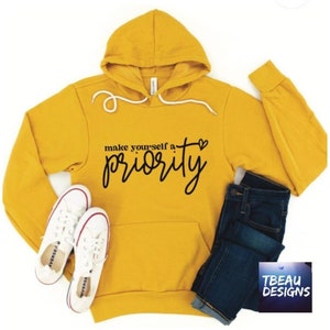 May include: Mustard yellow hoodie with the text "make yourself a priority" in black script. The hoodie has a drawstring hood and a front pocket. White sneakers and blue jeans are next to the hoodie.