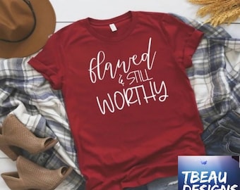 May include: A crimson t-shirt featuring the white text "Flawed & Still Worthy". The shirt is styled with a brown hat, a plaid scarf, and tan open-toe booties. The TBEAU DESIGNS logo is visible in the bottom right corner.