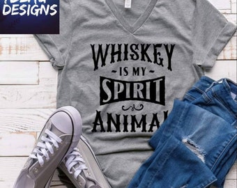 May include: A gray V-neck t-shirt with the text "Whiskey is my Spirit Animal" in black. The shirt is paired with blue jeans and gray sneakers with white laces. The image includes the logo "TBEAU DESIGNS".