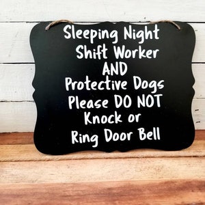 May include: Black chalkboard sign with white text: "Sleeping Night Shift Worker AND Protective Dogs Please DO NOT Knock or Ring Door Bell." The sign has a decorative shape and a twine hanger.