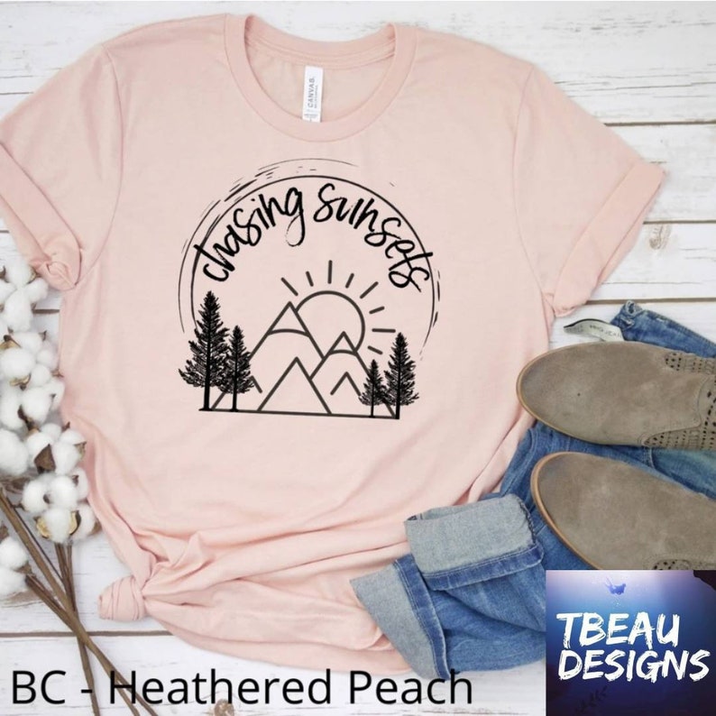 May include: A light pink t-shirt with rolled-up sleeves featuring a black graphic of mountains, trees, and a sun, encircled by the words "chasing sunsets." The shirt is paired with blue jeans and tan shoes.